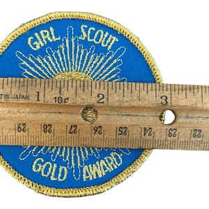 May include: A round Girl Scout Gold Award patch with blue fabric and gold embroidery. The patch is approximately 3 inches in diameter. The text "GIRL SCOUT GOLD AWARD" is embroidered in gold. A wooden ruler is placed over the patch.