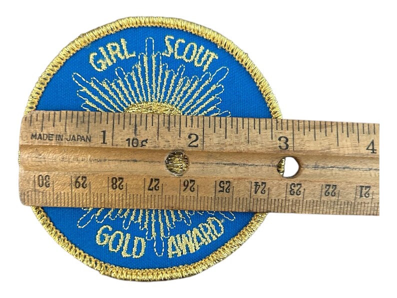 May include: A round Girl Scout Gold Award patch with blue fabric and gold embroidery. The patch is approximately 3 inches in diameter. The text "GIRL SCOUT GOLD AWARD" is embroidered in gold. A wooden ruler is placed over the patch.