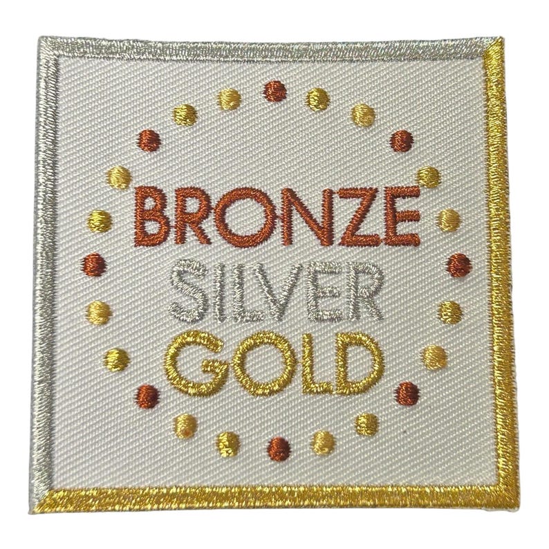 May include: Square embroidered patch with the words "BRONZE", "SILVER", and "GOLD" in metallic thread. The patch has a white background with a silver border and a gold border. Small dots in gold and bronze surround the words.