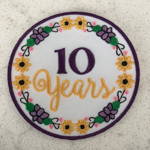 May include: A white embroidered patch with a purple 10 and gold lettering that says "years" surrounded by a wreath of yellow and purple flowers.