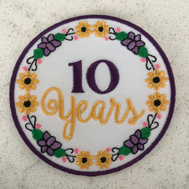 May include: A round, white embroidered patch with a purple border. The patch features the number "10" in purple and the word "Years" in gold script. A floral wreath of yellow sunflowers, purple flowers, and green leaves surrounds the text.