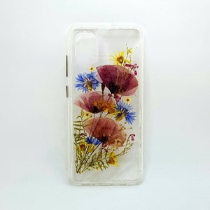 May include: A clear phone case featuring a pressed flower design. The flowers are pink, blue and yellow. The case is designed for a phone with a camera bump.