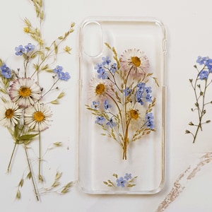May include: A clear phone case with pressed flowers and leaves. The flowers are white daisies and blue forget-me-nots. The case is designed for a smartphone.