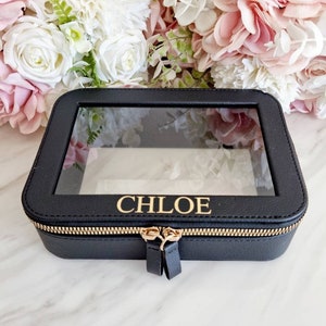 Puede incluir: Estuche de joyería rectangular negro con tapa transparente. El estuche tiene una cremallera dorada y el nombre "CHLOE" en letras doradas. El fondo presenta flores blancas y rosas.