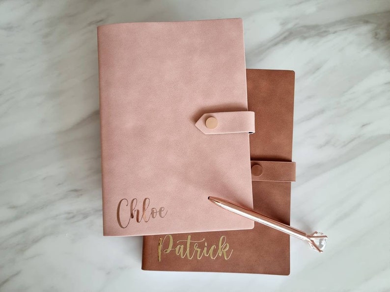 May include: Two personalized leather journals, one pink with the name "Chloe" and one brown with the name "Patrick", are stacked on top of each other. A rose gold pen with a diamond-shaped tip is lying on top of the journals.