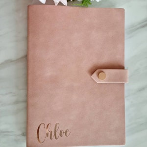 May include: A blush pink faux leather journal with a gold button closure and the name "Chloe" embossed in gold on the front cover.