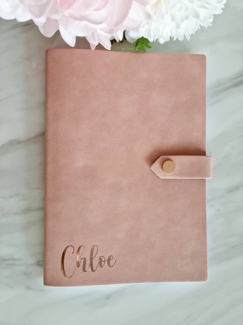 May include: A blush pink faux leather journal with a gold button closure and the name "Chloe" embossed in gold on the front cover.