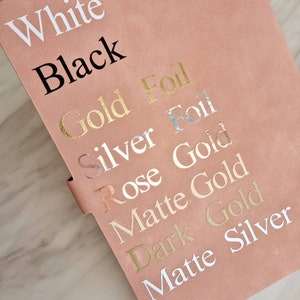May include: A pink notebook with the words "White", "Black", "Gold Foil", "Silver Foil", "Rose Gold", "Matte Gold", "Dark Gold", and "Matte Silver" written on it in different colors.