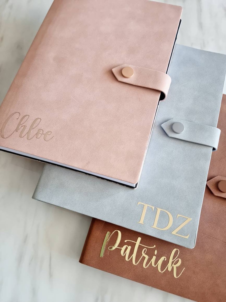 May include: Three personalized notebooks with gold lettering. The top notebook is pink with the name "Chloe" on the front. The middle notebook is gray with the initials "TDZ" and the name "Patrick" on the front. The bottom notebook is brown.