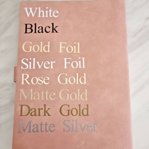 May include: A pink notebook with the words "White", "Black", "Gold Foil", "Silver Foil", "Rose Gold", "Matte Gold", "Dark Gold", and "Matte Silver" written on the front cover.