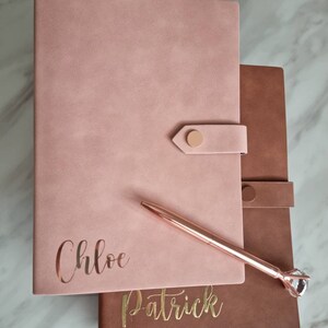 May include: Two personalized journals with gold lettering. The top journal is pink with the name "Chloe" and the bottom journal is brown with the name "Patrick". A rose gold pen with a crystal top is lying on top of the journals.