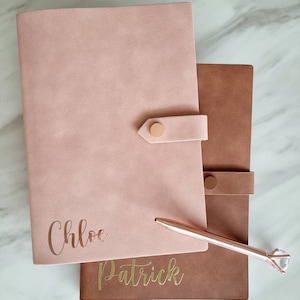 May include: Two personalized leather journals with gold lettering. The top journal is pink and says "Chloe". The bottom journal is brown and says "Patrick". A rose gold pen with a clear crystal top is lying on top of the journals.