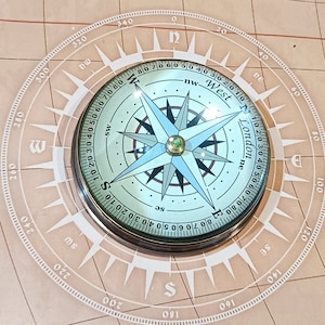 Może przedstawiać: Kompas w stylu vintage z szczegółową tarczą, z oznaczeniami kierunków i centralnym wzorem gwiazdy. Kompas jest umieszczony na tle mapy świata. Widoczny jest tekst "Crafts and Ingraving". Jest to narzędzie nawigacyjne.