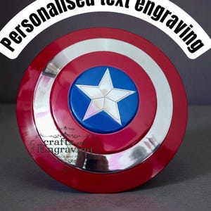 May include: A red, white, and blue shield with a silver rim and a white star on a blue center. The shield has the text "Personalised text engraving" in a white arch. The shield is a replica of a superhero's shield.