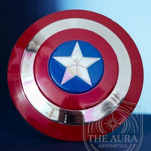 May include: A Captain America shield replica. The shield is circular, featuring a red outer ring, a silver inner ring, and a blue central disc with a silver star. The shield has a glossy finish and is set against a dark blue backdrop.
