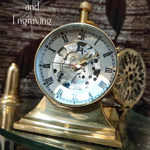 May include: A brass-colored, skeleton clock with a clear glass face, showcasing the intricate inner workings. The clock features Roman numerals and gold-colored hands. The base is also brass-colored, and the image includes the text "Crafts and Engraving".