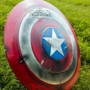 May include: A red, white, and blue Captain America shield with a distressed, weathered look. The shield is lying on a bed of green grass. The shield has a star in the center and concentric circles around the star. The shield has a small logo on the top that says "crafts and Engraving".