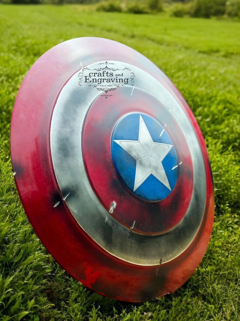 May include: A red, white, and blue Captain America shield with a distressed, weathered look. The shield is lying on a bed of green grass. The shield has a star in the center and concentric circles around the star. The shield has a small logo on the top that says "crafts and Engraving".