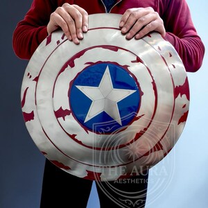 May include: A circular shield in silver, with a blue central disc and a white star. The shield has red markings around the edge. The text "THE AURA AESTHETICS" is visible. The shield is held by a person.