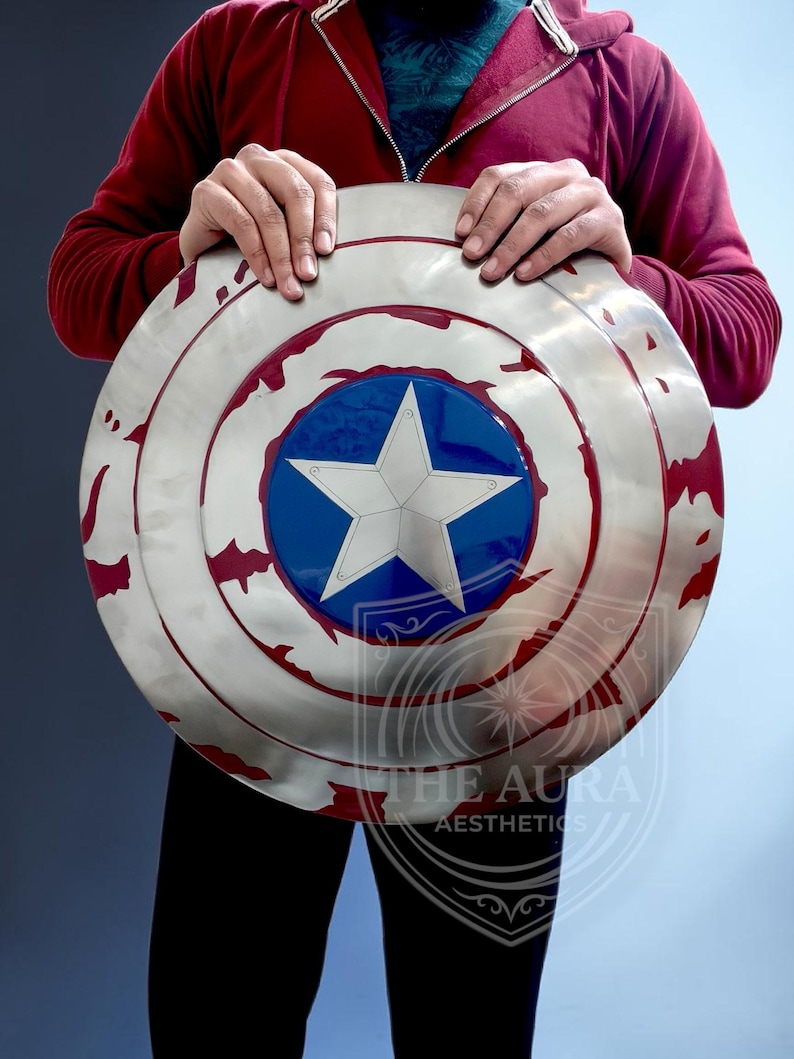 May include: A circular shield in silver, with a blue central disc and a white star. The shield has red markings around the edge. The text "THE AURA AESTHETICS" is visible. The shield is held by a person.