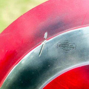 May include: A close-up of a red and silver metal disc with the words "crafts and engraving" in black text. The disc has a small dent in the silver part.