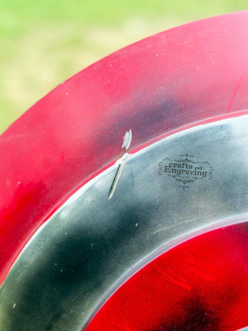 May include: A close-up of a red and silver metal disc with the words "crafts and engraving" in black text. The disc has a small dent in the silver part.