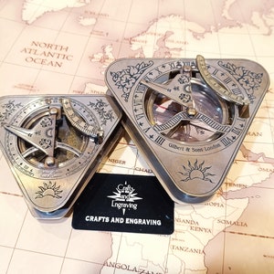 May include: Two antique brass sundial compasses with intricate designs and a compass needle. The compasses are triangular in shape and have a compass needle, a sundial, and a compass rose. The compasses are on a map with the text "Gilbert & Sons London" on one of the compasses.