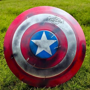 May include: A red, white, and blue Captain America shield with a distressed paint finish. The shield has a five-pointed star in the center and the words "crafts and Engraving" on the top.