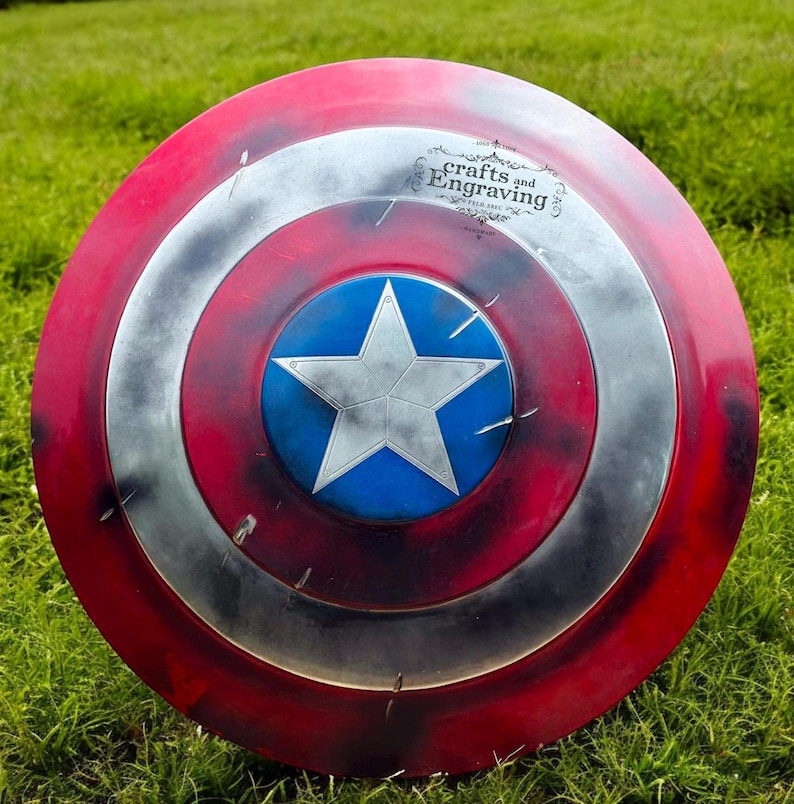 May include: A red, white, and blue Captain America shield with a distressed paint finish. The shield has a five-pointed star in the center and the words "crafts and Engraving" on the top.