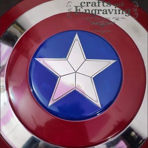 Puede incluir: Una r&eacute;plica del escudo del Capit&aacute;n Am&eacute;rica. El escudo presenta un dise&ntilde;o rojo, blanco y plateado con un disco central azul. Una estrella plateada de cinco puntas est&aacute; centrada en el disco azul. El escudo tiene el texto "crafts and engraving".