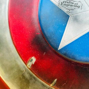 May include: A close-up of a red, white, and blue American shield with a silver star in the center. The shield is made of metal and has a distressed, weathered look. The text "Crafts and Engraving" is printed on the star.