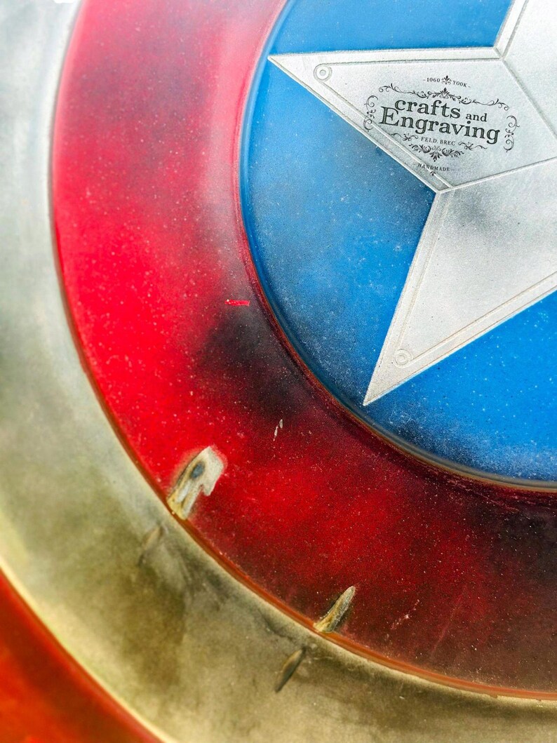 May include: A close-up of a red, white, and blue American shield with a silver star in the center. The shield is made of metal and has a distressed, weathered look. The text "Crafts and Engraving" is printed on the star.
