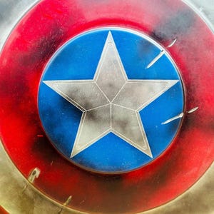May include: A close-up of a red, white, and blue Captain America shield with a large, silver star in the center. The shield has a distressed, weathered look, with visible scratches and paint chips.