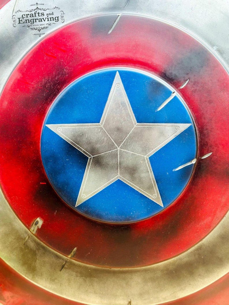 May include: A close-up of a red, white, and blue Captain America shield with a large, silver star in the center. The shield has a distressed, weathered look, with visible scratches and paint chips.