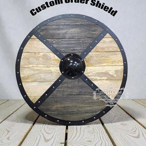 Pode incluir: Um escudo de madeira redondo com aro e detalhes em metal preto. O escudo tem um chefe central preto e a inscrição "Custom Order Shield". A superfície de madeira é dividida em seções de madeira clara e escura.