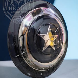 May include: A black, circular shield with a silver star at its centre and a silver border. The shield has a reflective surface and is set against a blue and white backdrop. The text "THE AURA AESTHETICS" is visible in the upper left corner.