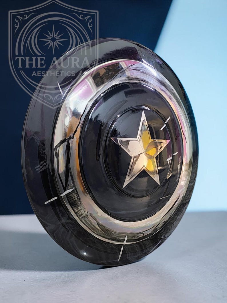 May include: A black, circular shield with a silver star at its centre and a silver border. The shield has a reflective surface and is set against a blue and white backdrop. The text "THE AURA AESTHETICS" is visible in the upper left corner.
