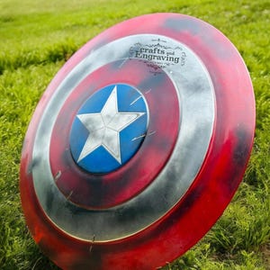 May include: A red, white, and blue Captain America shield with a star in the center. The shield is lying on a bed of green grass. The shield has a distressed look with some paint chipping and scratches. The shield has the text "crafts and Engraving" on the top.