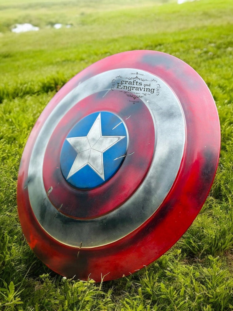 May include: A red, white, and blue Captain America shield with a star in the center. The shield is lying on a bed of green grass. The shield has a distressed look with some paint chipping and scratches. The shield has the text "crafts and Engraving" on the top.