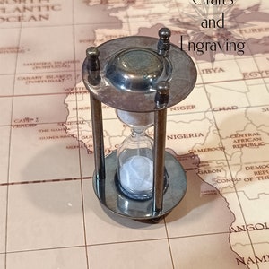 May include: A brass hourglass with a white sand timer on a world map background. The hourglass is on a wooden surface. The text "Crafts and Engraving" is visible in the upper right corner of the image.