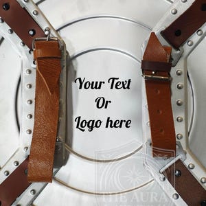 May include: A silver shield with brown leather straps and buckles. The shield has a circular design with rivets and the text "Your Text Or Logo here". The Aura Aesthetics logo is at the bottom.