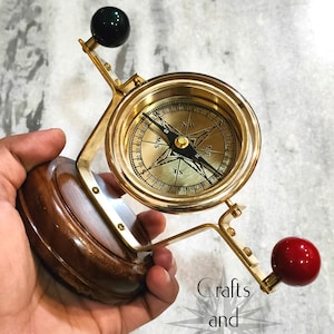 May include: A brass compass mounted on a wooden base, featuring a rotating dial and a needle pointing north. The compass has a green and a red sphere on each side. The text "Crafts and Engraving" is visible.