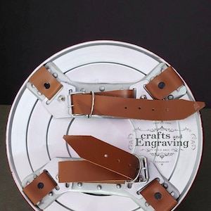 Puede incluir: Un escudo hecho a mano con un borde rojo y una cara blanca con c&iacute;rculos conc&eacute;ntricos. La parte trasera tiene correas de cuero marr&oacute;n y soportes de metal plateado. El escudo tiene el texto "crafts and Engraving" y "HANDMADE".