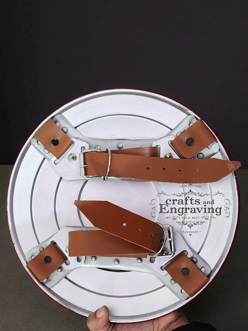 Puede incluir: Un escudo hecho a mano con un borde rojo y una cara blanca con c&iacute;rculos conc&eacute;ntricos. La parte trasera tiene correas de cuero marr&oacute;n y soportes de metal plateado. El escudo tiene el texto "crafts and Engraving" y "HANDMADE".