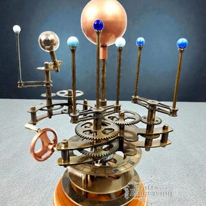 May include: A brass and wood orrery model of the solar system. The model features a large copper sphere representing the sun, with smaller spheres and gears representing the planets. The base is made of wood, and the model has a hand crank.