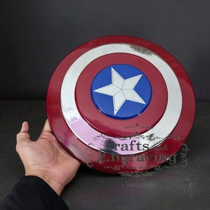 Puede incluir: Un escudo del Capit&aacute;n Am&eacute;rica rojo, blanco y azul con un emblema de estrella. El escudo tiene un dise&ntilde;o circular con un borde plateado y el texto "crafts and engraving".