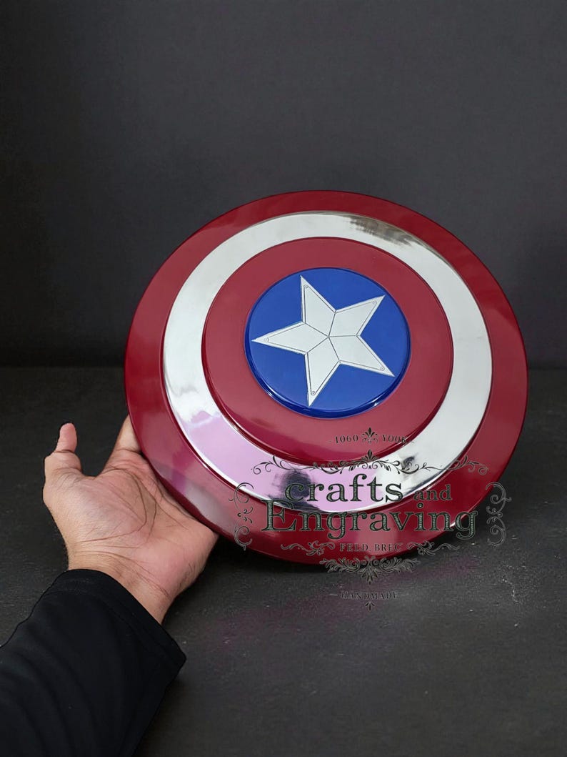 Puede incluir: Un escudo del Capit&aacute;n Am&eacute;rica rojo, blanco y azul con un emblema de estrella. El escudo tiene un dise&ntilde;o circular con un borde plateado y el texto "crafts and engraving".