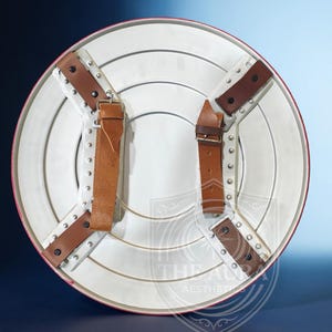 May include: A white and red shield with brown leather straps and metal rivets. The shield has a circular design with concentric rings and a red border. The back features four leather straps for securing the shield.