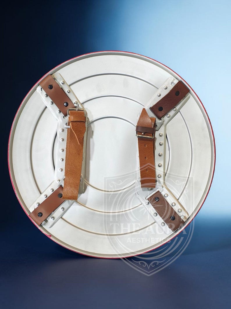May include: A white and red shield with brown leather straps and metal rivets. The shield has a circular design with concentric rings and a red border. The back features four leather straps for securing the shield.