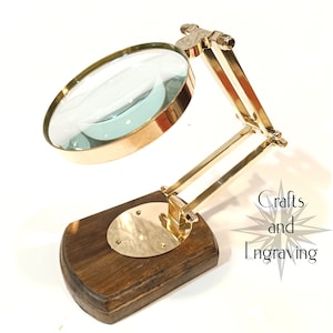 May include: A brass magnifying glass with a wooden base. The magnifying glass is on a stand and can be adjusted to different angles. The stand is made of dark wood and has a brass plate on the bottom.